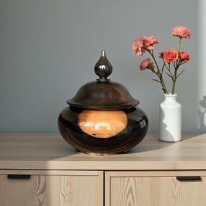 Pode incluir: Um pote decorativo de madeira castanha escura com tampa e um pequeno remate arredondado. O pote tem um acabamento brilhante e um interior de cor clara. À direita, um pequeno vaso branco com flores cor-de-rosa.