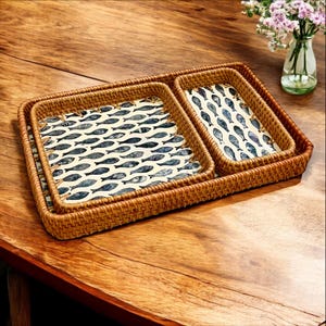May include: Two rectangular woven rattan trays with a fish pattern. The fish are blue and grey on a cream background. The trays are on a wooden table. The trays are ideal for serving food.