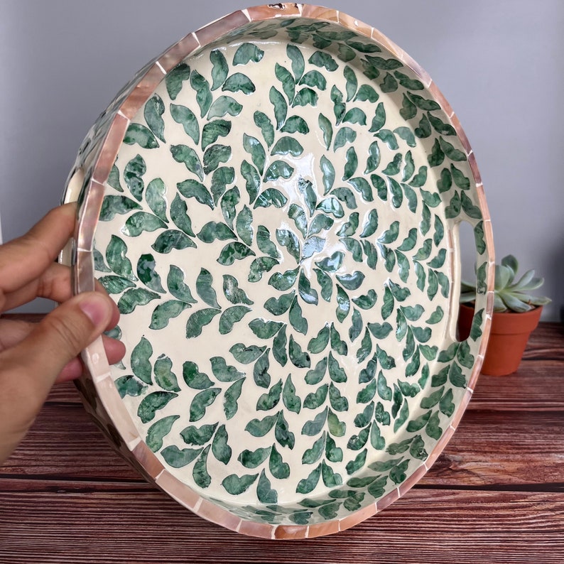 Op de afbeelding: Een ronde dienblad met een witte achtergrond en een groen bladmotief. Het dienblad heeft aan elke kant een handvat en is gemaakt van parelmoer.