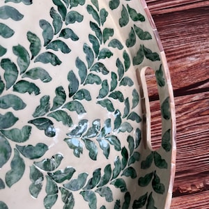 Op de afbeelding: Een ronde, witte dienblad met een groen bladmotief. Het dienblad heeft twee handvatten aan de zijkant.
