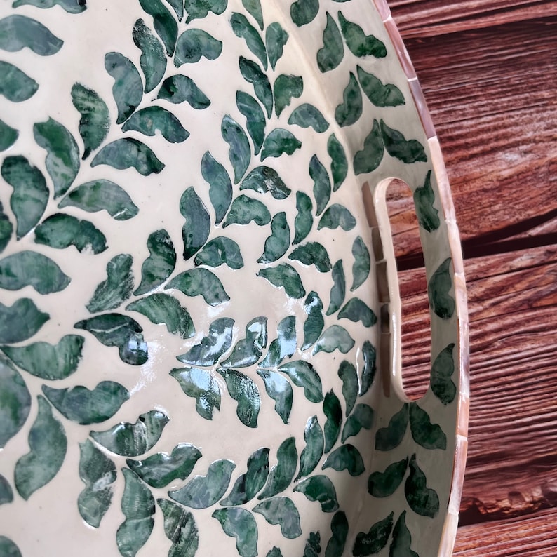 Op de afbeelding: Een ronde, witte dienblad met een groen bladmotief. Het dienblad heeft twee handvatten aan de zijkant.