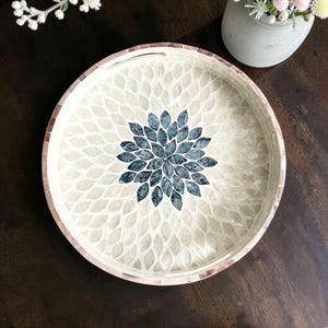May include: Round decorative serving tray with a mosaic design. The tray features a central floral pattern in shades of blue and a border of light-coloured tiles. The outer rim is a mosaic of light and dark brown tiles. The tray is approximately 30 cm in diameter.