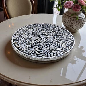 May include: A circular decorative tray featuring a black base and a leaf design crafted from white and silver inlays. The tray rests on a cream-coloured table. A white vase with pink roses is also visible.