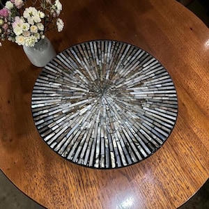 May include: A circular decorative tray featuring a sunburst pattern composed of iridescent, rectangular mother-of-pearl tiles in shades of gray, silver, and white, arranged in a radiating design from the center to the outer black rim.