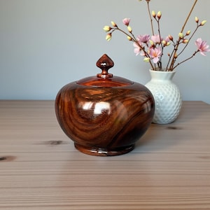 Op de afbeelding: Een handgemaakte houten pot met een deksel, met een rijk, wervelend nerfpatroon in tinten bruin en roodbruin. De pot heeft een afgeronde vorm en een kleine, puntige bekroning op het deksel. Een witte vaas met roze bloemen staat op de achtergrond.