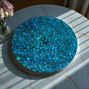 Op de afbeelding: Een ronde, decoratieve schaal met een mozaïekontwerp in tinten van blauwgroen en blauw. Het mozaïkpatroon lijkt op visschubben. De schaal staat op een witte tafel met marmerlook, met een vaas met roze en witte bloemen op de achtergrond.
