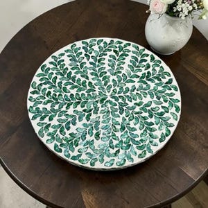 May include: A round, white decorative tray with a mosaic leaf pattern in shades of green. The tray is placed on a dark brown wooden table. A white vase with pink and white flowers is in the background.