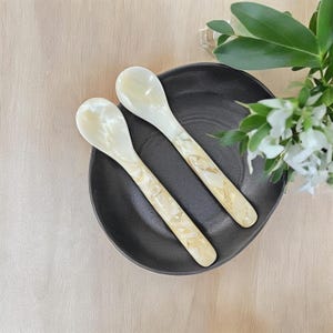 May include: Two white mother of pearl spoons with a swirl pattern on a black plate.