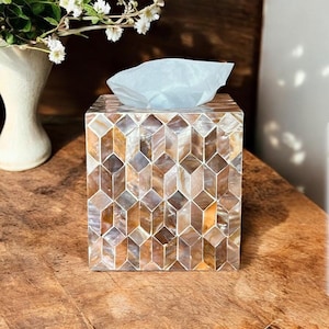 May include: A square tissue box with a geometric pattern of iridescent shell pieces in shades of brown, tan, and cream. A white tissue is partially pulled from the top. The box sits on a wooden surface.