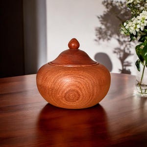 May include: A wooden lidded jar with a rounded body and a smooth, polished finish. The jar is made of reddish-brown wood with a natural grain pattern.