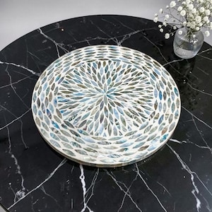 Op de afbeelding: Een rond decoratief dienblad met een ingewikkeld mozaïekpatroon van iriserende schelpstukjes in tinten wit, blauw en grijs. De individuele schelpsegmenten zijn gerangschikt in een radiaal, bloembladachtig ontwerp over het gehele oppervlak.