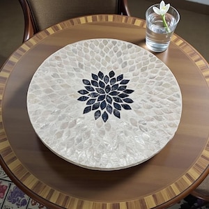 Pode incluir: Uma bandeja decorativa redonda com um design de mosaico de madrepérola. A bandeja apresenta um padrão floral central em azul escuro, rodeado por um padrão branco e iridescente em forma de folha. Está sobre uma mesa de madeira, ao lado de um copo de água com uma flor.