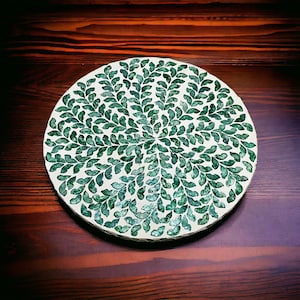 May include: A round, white coaster with a green leaf design. The coaster is made of a mosaic of small, green pieces.