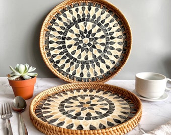 May include: Two round decorative trays with woven rattan edges. The tray centers feature a mosaic pattern in black, cream, and beige. One tray is in the foreground, and the other is slightly behind it. The trays are on a marble surface.