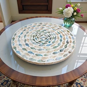 Op de afbeelding: Een ronde decoratieve schaal met een vismozaïekontwerp. Het mozaïek bevat talrijke kleine vissen in tinten blauw, beige en wit, gerangschikt in concentrische cirkels. De schaal staat op een ronde tafel met een glazen blad.