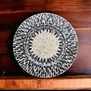 May include: A round decorative plate with a black and white mosaic design. The center of the plate features a white flower-like pattern.