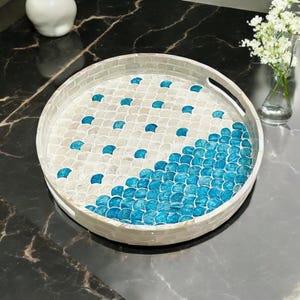 May include: A round tray with a white and blue mosaic design. The tray is made of mother of pearl and features a scalloped edge.