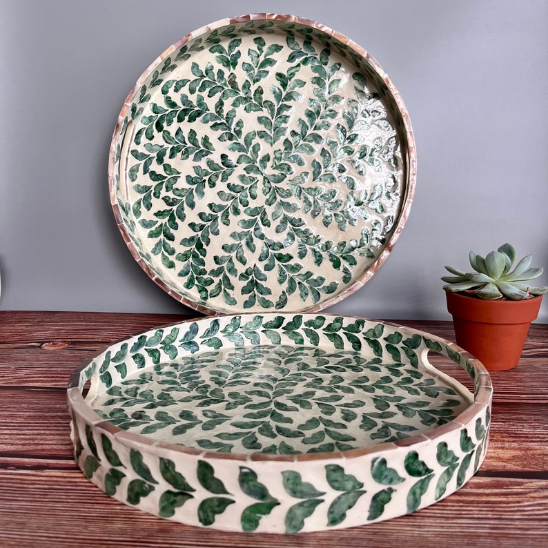 Op de afbeelding: Twee ronde schalen met een witte achtergrond en een groen bladmotief. De grotere schaal ligt bovenop de kleinere. De kleinere schaal heeft aan elke kant een handvat.