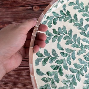 Op de afbeelding: Een ronde dienblad met een witte achtergrond en een groen bladmotief. Het dienblad is gemaakt van parelmoer en heeft een verhoogde rand.