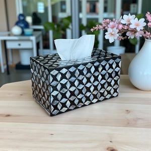 May include: A decorative tissue box with a black and white starburst pattern. The box is rectangular and has a slot at the top for tissue dispensing. A small bouquet of pink flowers in a white vase sits nearby.