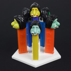 May include: A set of six colorful plastic toy figures with different facial expressions. The figures are arranged in a circle on a white hexagonal base. Each figure has a different colored body and a black hairpiece.