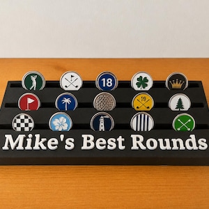 Op de afbeelding: Een zwarte rechthoekige displaystandaard met drie horizontale sleuven met vijftien ronde golfbalmarkers. De basis bevat de witte tekst Mike's Best Rounds. De markers tonen diverse iconen waaronder een golfer, vlaggen, een kroon en een vuurtoren.