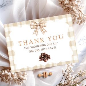 May include: A baby shower thank you card with a beige gingham border and a scalloped white center. The card features a watercolor bow, coffee beans, and the text "THANK YOU FOR SHOWERING OUR LIL' TINI ONE WITH LOVE!" A wooden rattle is at the bottom.