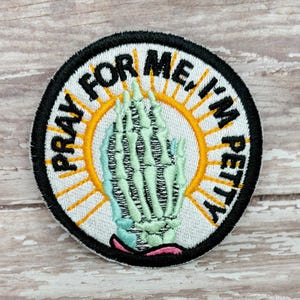 May include: A white embroidered patch with a black outline. The patch features a skeleton hand in a praying position with a yellow sun behind it. The text "Pray for me I'm petty" is written around the outside of the patch.