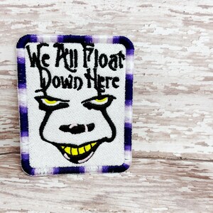 Puede incluir: Parche bordado con el texto "We All Float Down Here" y una cara de dibujos animados con ojos amarillos y una sonrisa amarilla. El parche tiene un fondo blanco con un borde a cuadros morado y blanco.