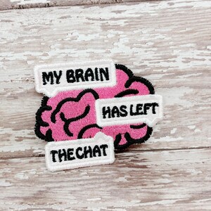 May include: A pink brain-shaped patch with black stitching and the text "MY BRAIN HAS LEFT THE CHAT" in white speech bubbles. The patch is on a light wood-grain surface.