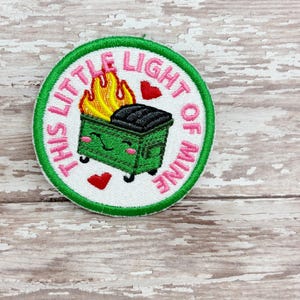 May include: Embroidered patch with a green border and the text "THIS LITTLE LIGHT OF MINE" in pink. The patch features a green dumpster with flames and hearts.