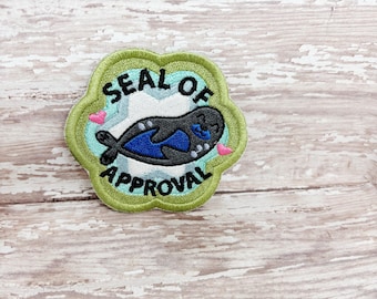 May include: Embroidered patch featuring a cartoon seal with a blue body and black flippers. The seal is inside a green, flower-shaped border with the words "SEAL OF APPROVAL" in black. Small pink hearts are on either side of the seal.