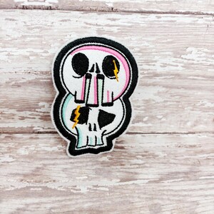 May include: Embroidered patch with a stacked skull design. The top skull has a pink outline, while the bottom skull has a teal outline. Both skulls have black eye sockets and a yellow lightning bolt accent. The patch is outlined in black.