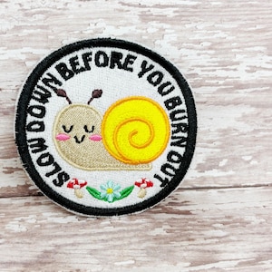 Può includere: Toppa ricamata con sfondo bianco e bordo nero. La toppa presenta una lumaca dei cartoni animati con un guscio giallo e la scritta "SLOW DOWN BEFORE YOU BURN OUT". Piccoli funghi e un fiore in basso.