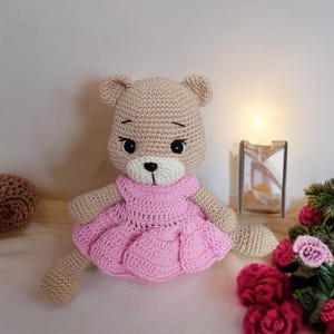 Può includere: Un orsacchiotto di peluche all'uncinetto che indossa un vestito rosa. L'orso ha gli occhi neri e un naso bianco. L'orso è seduto su una superficie bianca con fiori rosa sullo sfondo.
