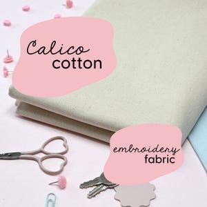 Könnte beinhalten: Ein Stapel beigefarbener Calico-Baumwollstoffe mit dem Text "Calico cotton" in einer rosa Form. Schere, Schlüssel und Reißzwecken sind um den Stoff herum verstreut. Der Text "embroidery fabric" ist ebenfalls sichtbar.