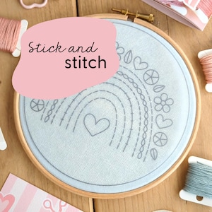 Puede incluir: Un aro de bordado azul claro con un diseño de arcoíris gris y el texto "Stick and stitch". El aro está rodeado de hilo de bordar de varios colores.