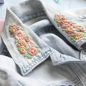 Könnte beinhalten: Eine hellblaue Jeansjacke mit detaillierter Blumenstickerei auf dem Kragen. Die Stickerei besteht aus rosa, pfirsichfarbenen und gelben Blüten mit grünen Blättern, die direkt auf den hellblauen Stoff der Kragenspitzen gestickt sind.