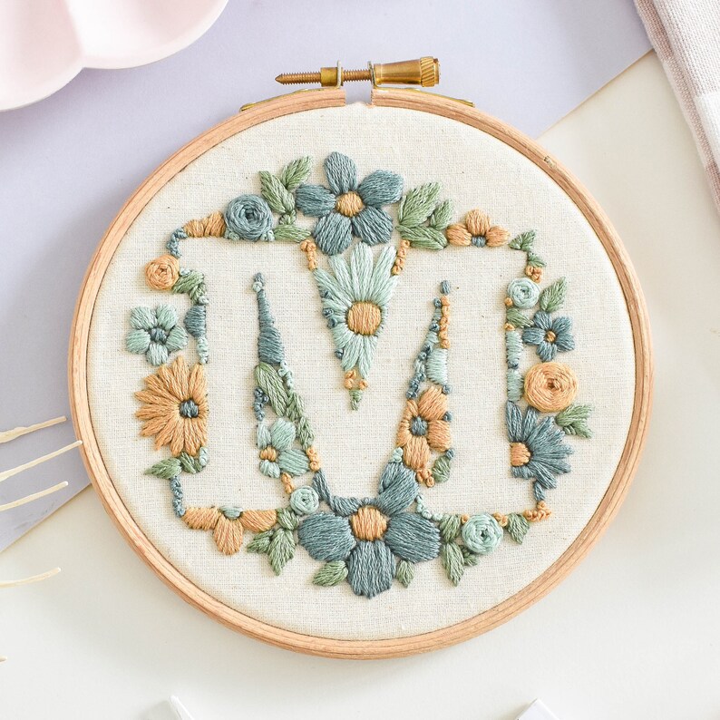 以下が含まれることがあります： 白い布地に緑、青、黄色の花柄が刺繍された円形の刺繍枠。デザインは、花に囲まれたスタイリッシュな文字「M」です。