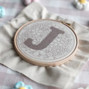 以下が含まれることがあります： 白い布地に花柄が刺繍された、茶色のアルファベット「J」が刺繍されています。布は木製の刺繍枠に張られています。