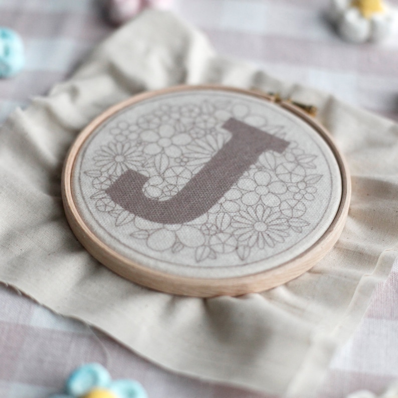 以下が含まれることがあります： 白い布地に花柄が刺繍された、茶色のアルファベット「J」が刺繍されています。布は木製の刺繍枠に張られています。