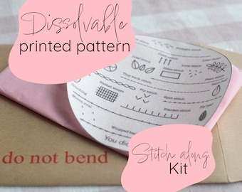 Puede incluir: Un sobre rosa con el texto "Dissolvable printed pattern" y "Stitch along Kit" en él. El sobre contiene un círculo de papel blanco con varios puntos de bordado dibujados en él.