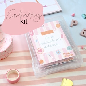 Puede incluir: Una caja de plástico transparente llena de suministros de bordado. La caja tiene un fondo a rayas rosa y blanco con el texto "One stitch at a time..." y una cara de dibujos animados linda. La caja está etiquetada como "Pick & Stitch".