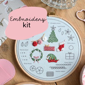 May include: An embroidery kit with a light blue fabric featuring various stitched designs, including a Christmas tree, wreath, and gift box. The kit includes a wooden hoop and the text "Embroidery kit" in a pink bubble.