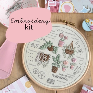 Pode incluir: Um kit de bordado com um bastidor de madeira com um design botânico. O design inclui plantas bordadas, flores e exemplos de pontos. O kit inclui agulhas, linha rosa e um porta-agulhas rosa.