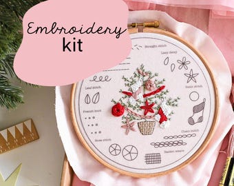 Peut inclure: Un kit de broderie avec un motif de sapin de Noël. Le kit comprend un cercle à broder en bois, du tissu avec un motif pré-imprimé et des fils à broder. Le texte "Embroidery kit" est affiché sur un fond rose. Le kit est emballé dans une boîte rose.