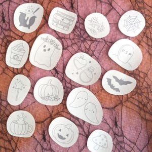 以下が含まれることがあります： ハロウィーンをテーマにした様々なイラストが描かれた、白い丸いステッカー15枚セット。イラストには、大釜、本、葉っぱ、蜘蛛の巣、コーヒーカップ、幽霊、魔法薬の瓶、墓石、コウモリ、カボチャ、魔法の杖、キノコ、マグカップなどが含まれています。