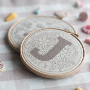 以下が含まれることがあります： 薄い茶色の刺繍枠のクローズアップ。枠の中にはベージュの麻布があり、花柄のデザインと中央に大きな濃い茶色の「J」の文字が描かれています。