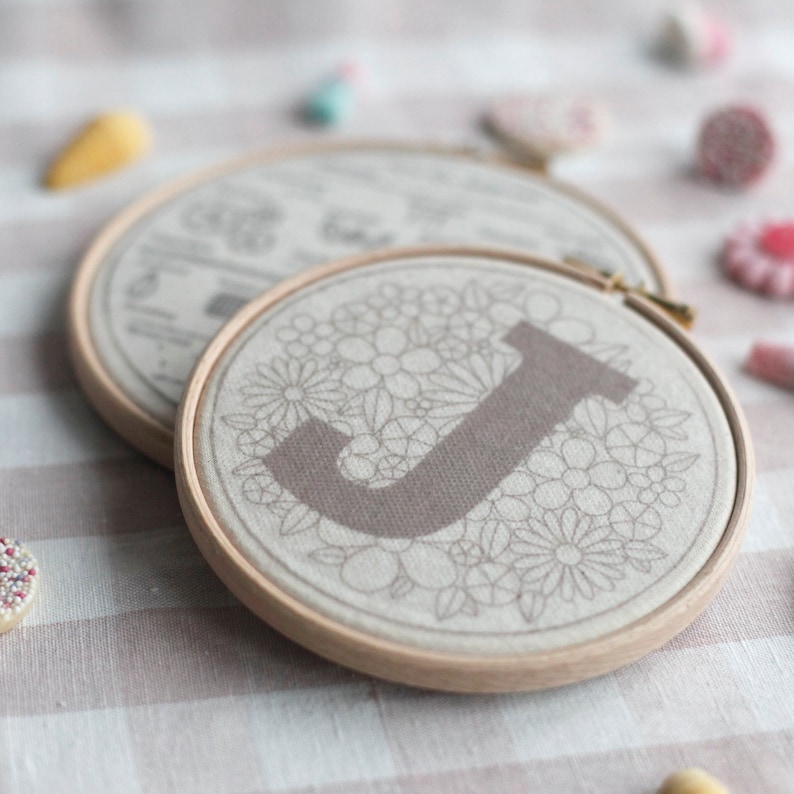 以下が含まれることがあります： 薄い茶色の刺繍枠のクローズアップ。枠の中にはベージュの麻布があり、花柄のデザインと中央に大きな濃い茶色の「J」の文字が描かれています。