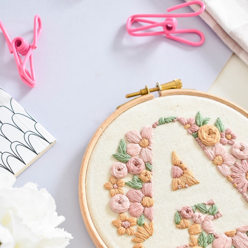以下が含まれることがあります： 白い布地に花柄の刺繍が施されたAの文字が描かれた、木製の刺繍枠。花はピンク、ピーチ、イエローです。